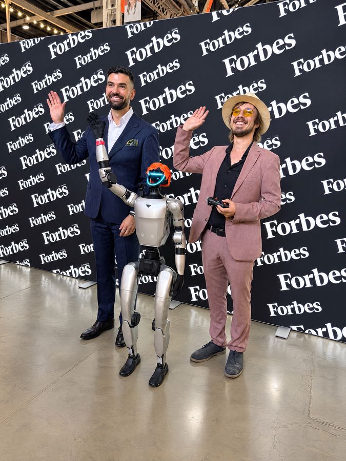Humanoidný robot Unitree G1 s oranžovou parochňou máva spolu s dvoma mužmi pred tlačovou stenou Forbes Slovensko, jeden z nich drží ovládač