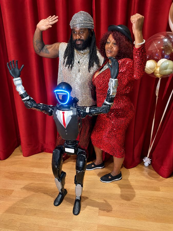 Humanoidný robot so zdvihnutými rukami pózuje s dvojicou hostí pred červeným závesom vo fotokútiku