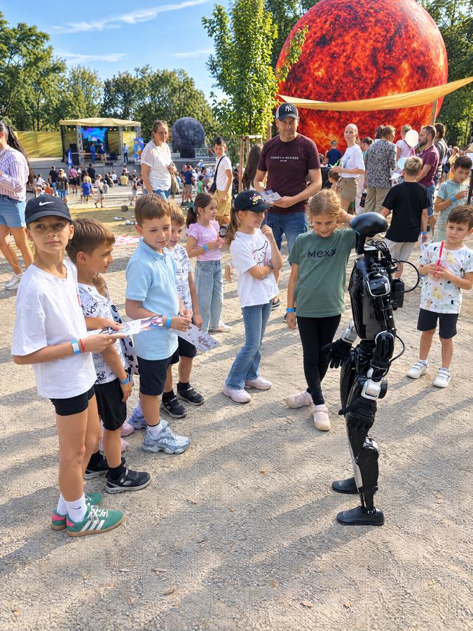 Humanoidný robot Unitree G1 stojí medzi deťmi na letnom festivale, v pozadí veľká socha planéty