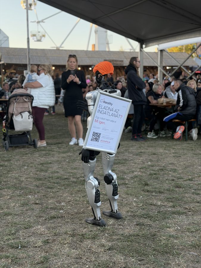 Humanoid robot hátulról, a hátán PlusReality reklámtáblával és QR-kóddal a vásár látogatói között, mögötte óriáskerék