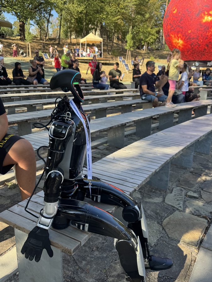 Humanoid robot sitting on a bench in the amphitheatre audience among festival spectators