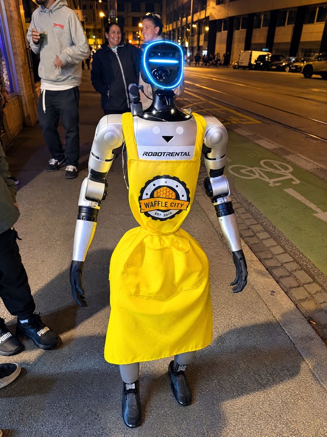 Humanoidný robot vo žltej zástere s logom Waffle City a so svetielkami večer na chodníku, okolo ľudia