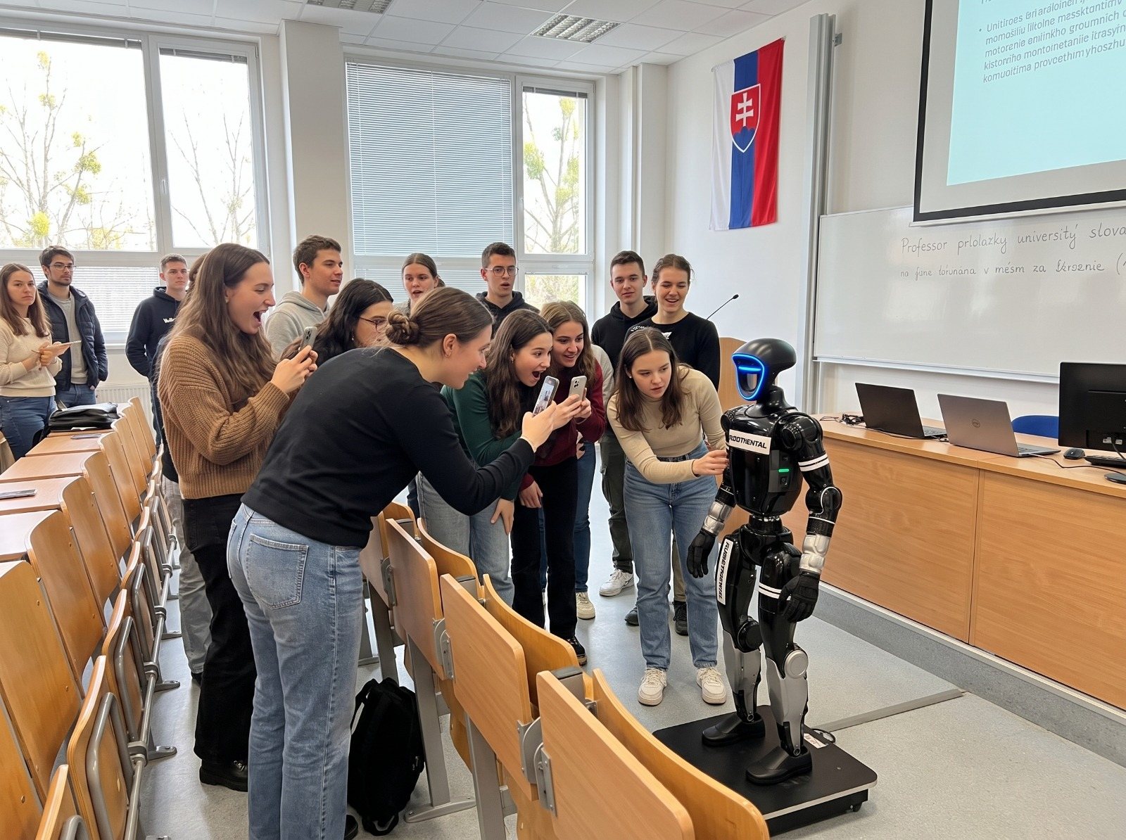 Estudiantes en un aula magna se inclinan hacia un robot humanoide sobre una base y lo fotografían