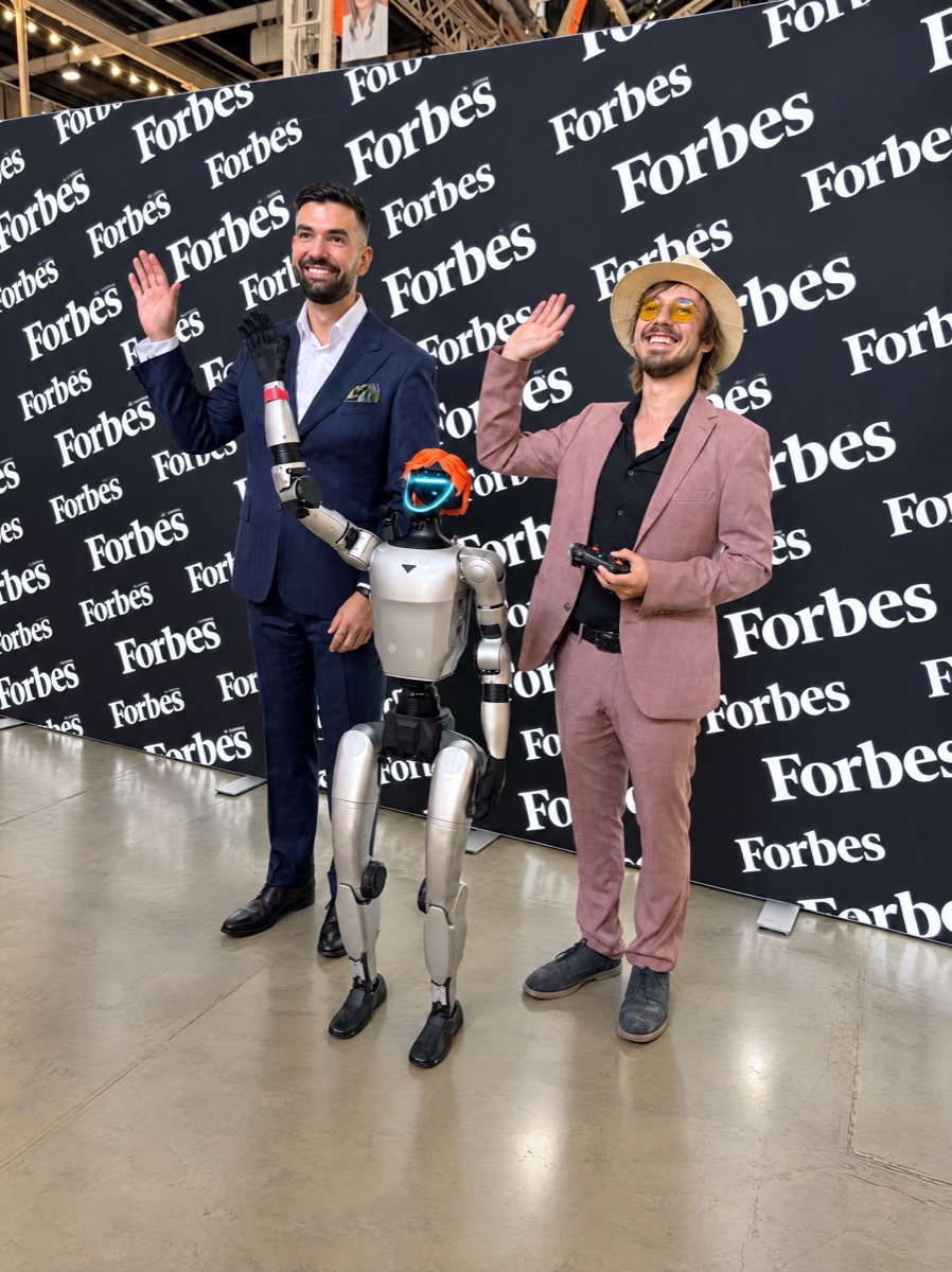 A humanoid robot in an orange wig waves together with two men in front of a Forbes press wall, one of them holding a controller