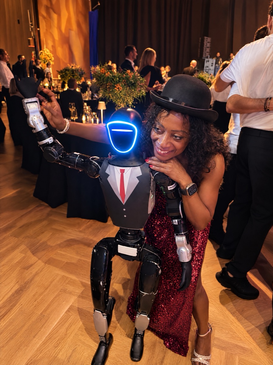 A guest at a gala dinner poses for a photo with a humanoid robot in a suit, red tie and hat, with laid tables and dancing guests behind them
