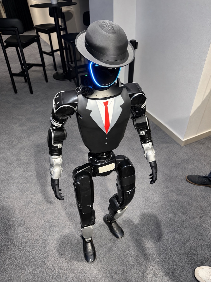 A Unitree G1 humanoid robot in a black suit, red tie and hat stands in the hall before the evening begins